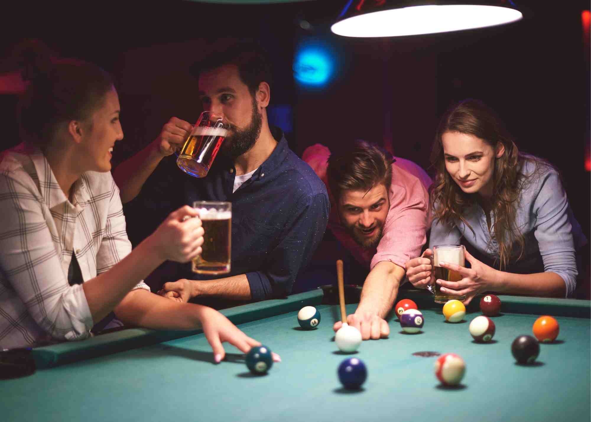 A group of four friends stand around a pool table, enjoying games and drinking beer.