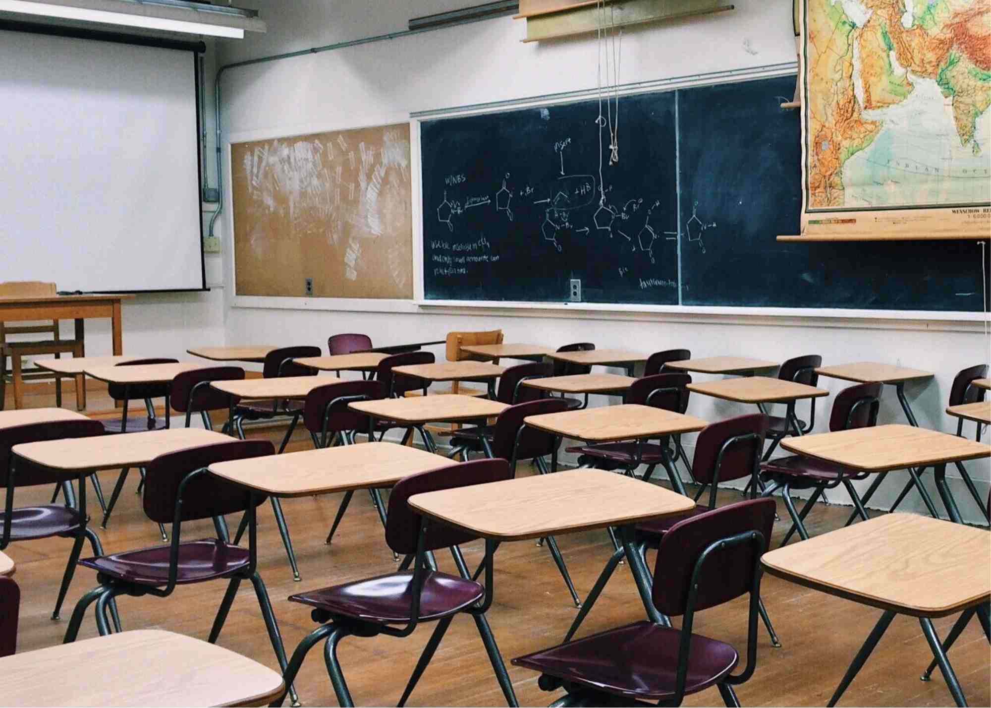 An empty classroom lined with desks. Along the walls are chalkboards, maps, and a projection screen.