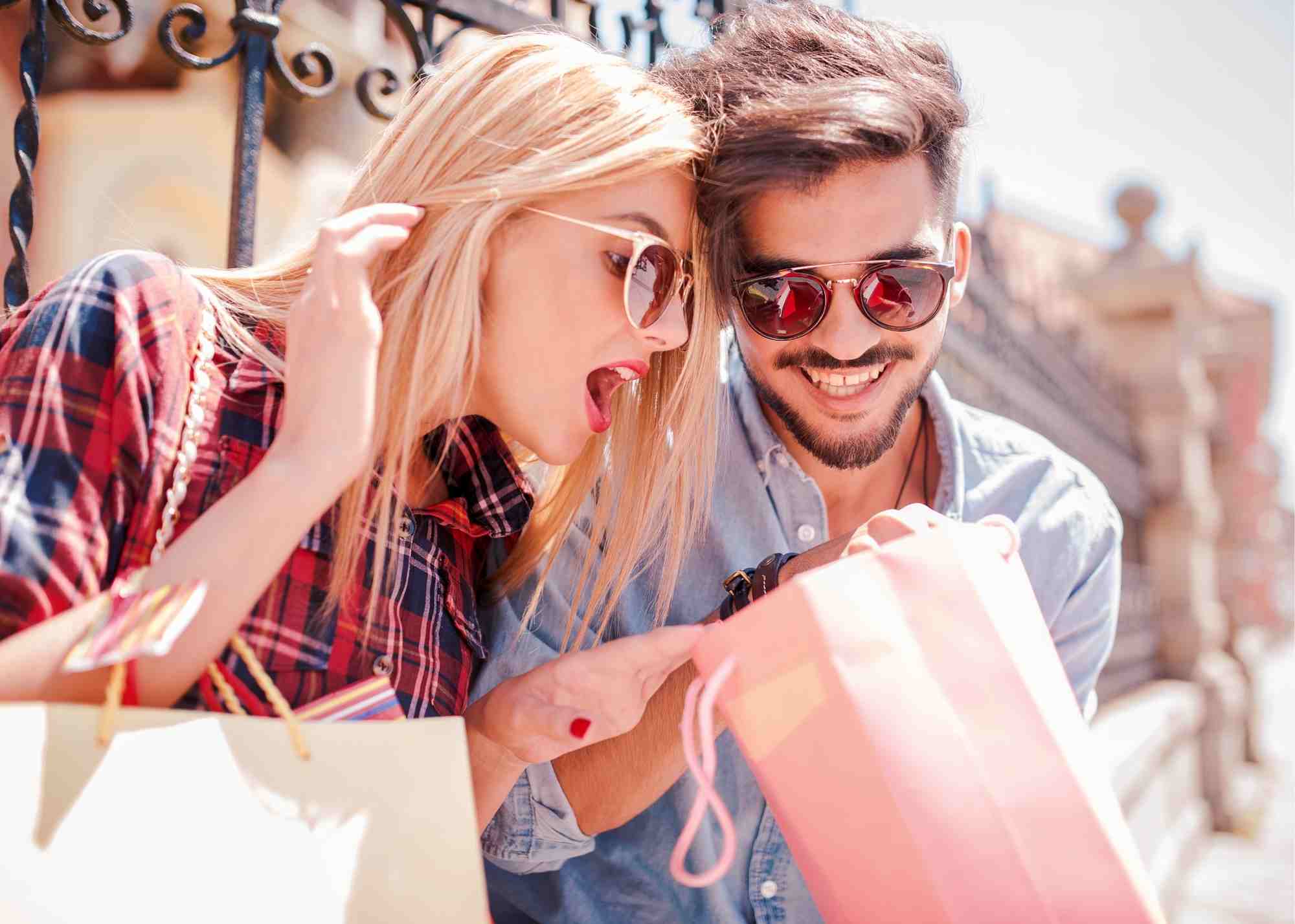 On a bright sunny day, a woman with a shopping bag, peaks into her friends bag with a gasp and smile.