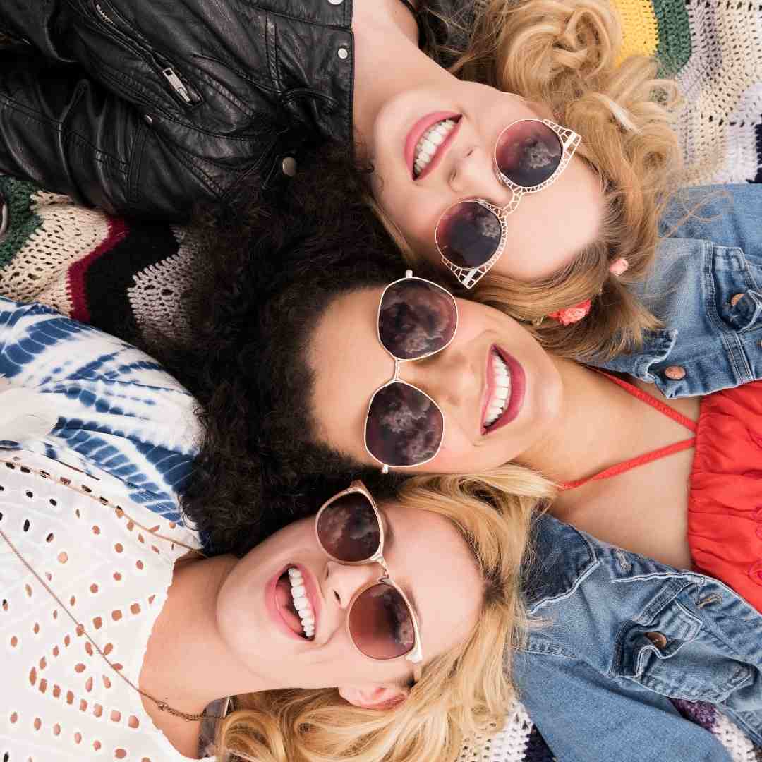 Three girls wearing sunglasses and smiles, lay beside each other, looking up.